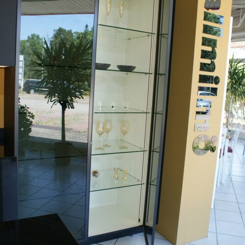 Geöffneter Vitrinenschrank mit Gläsern und Spiegel, Pflanzen im Hintergrund.