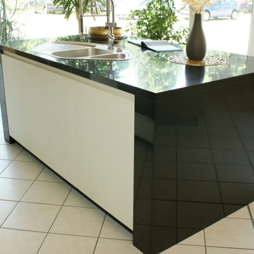 Schwarze Kücheninsel mit Spüle, glänzendem Finish, Dekovase und Pflanzen im Hintergrund.